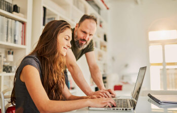 Eine Jugendliche sitzt lächelnd vor einem Laptop. Ein Mann schaut ihr dabei über die Schulter. 